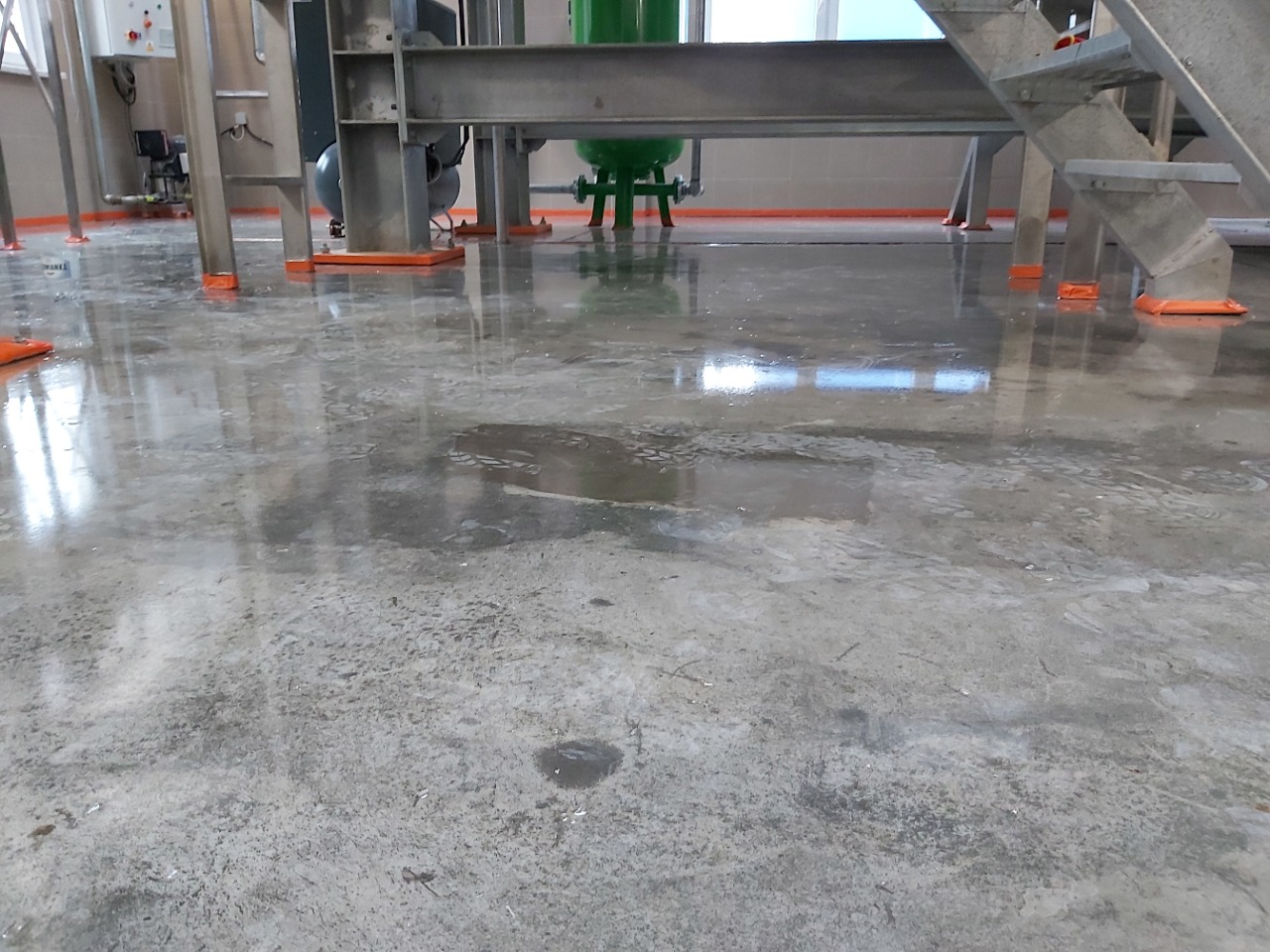 Błyszcząca, szara posadzka w hali przemysłowej, z widocznymi elementami konstrukcyjnymi i odblaskami światła.