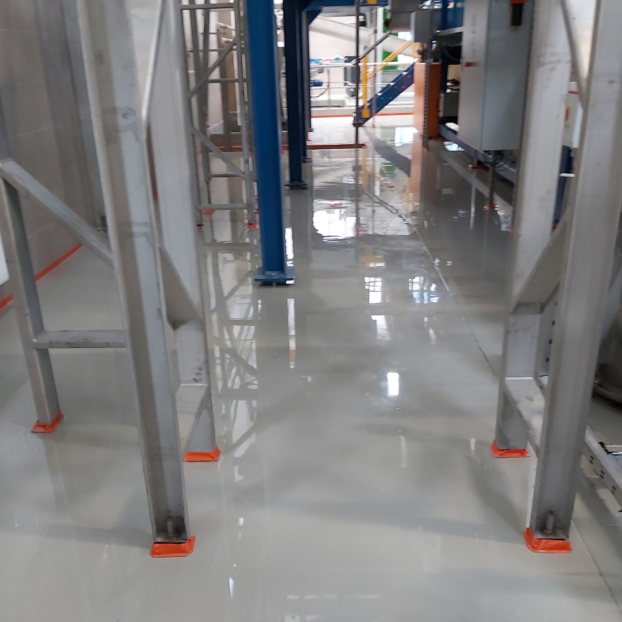 Jasnoszara, błyszcząca posadzka w hali przemysłowej, z metalowymi elementami konstrukcyjnymi i pomarańczowymi podkładkami pod nogami. Widoczne odbicia w posadzce.