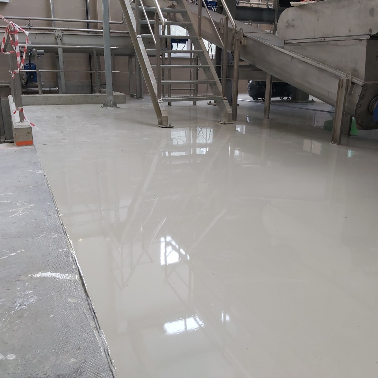 Błyszcząca, jasnoszara posadzka w hali przemysłowej, z widocznymi elementami metalowej konstrukcji i schodów.