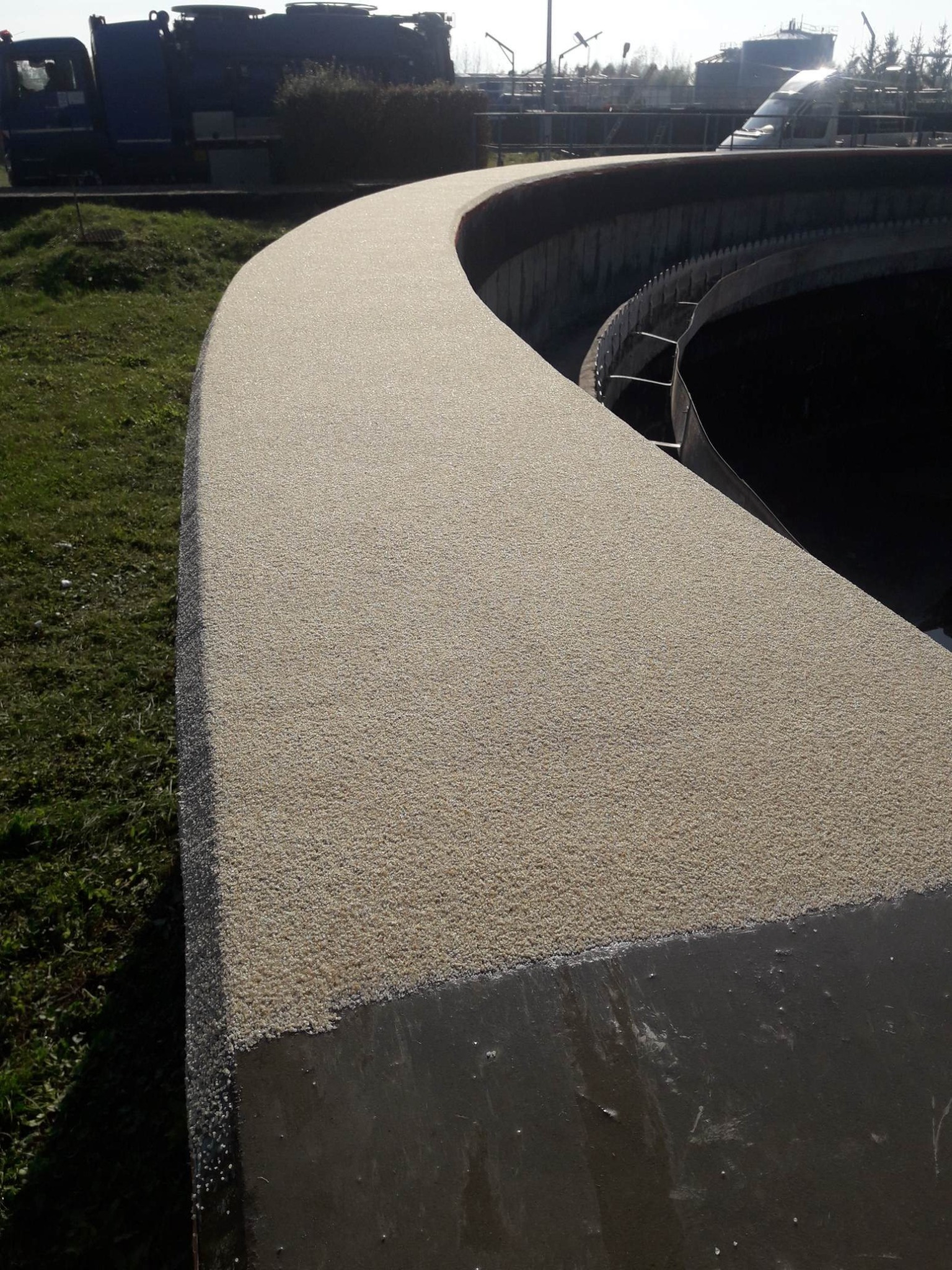 Jasna, drobnoziarnista powierzchnia na betonowej konstrukcji o zakrzywionym kształcie, prawdopodobnie element oczyszczalni ścieków, z widocznym fragmentem trawnika i pojazdu w tle.