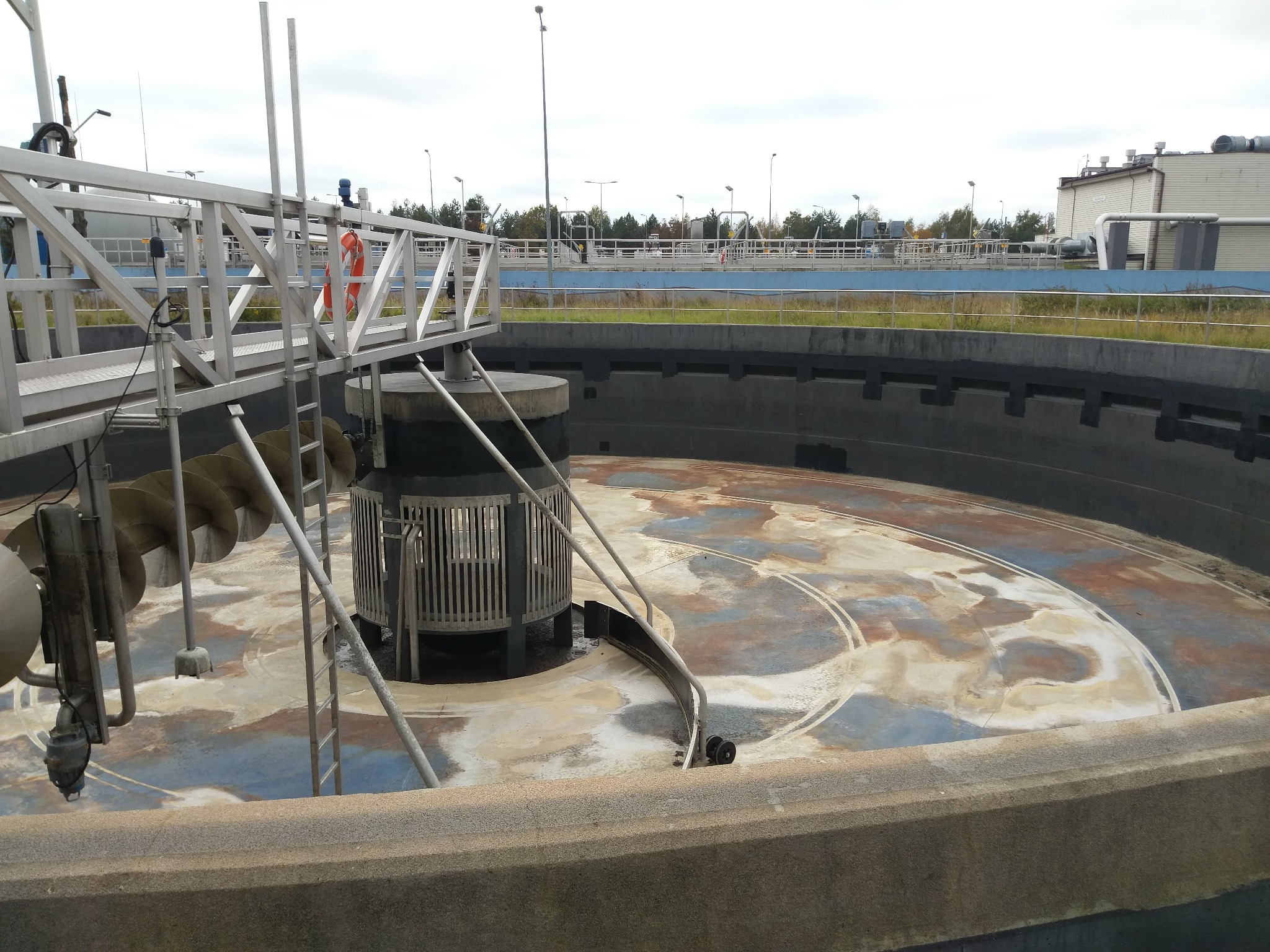 Oczyszczalnia ścieków z metalową konstrukcją pomostową i spiralnym podajnikiem, widok na okrągły zbiornik z osadem i śladami po czyszczeniu.