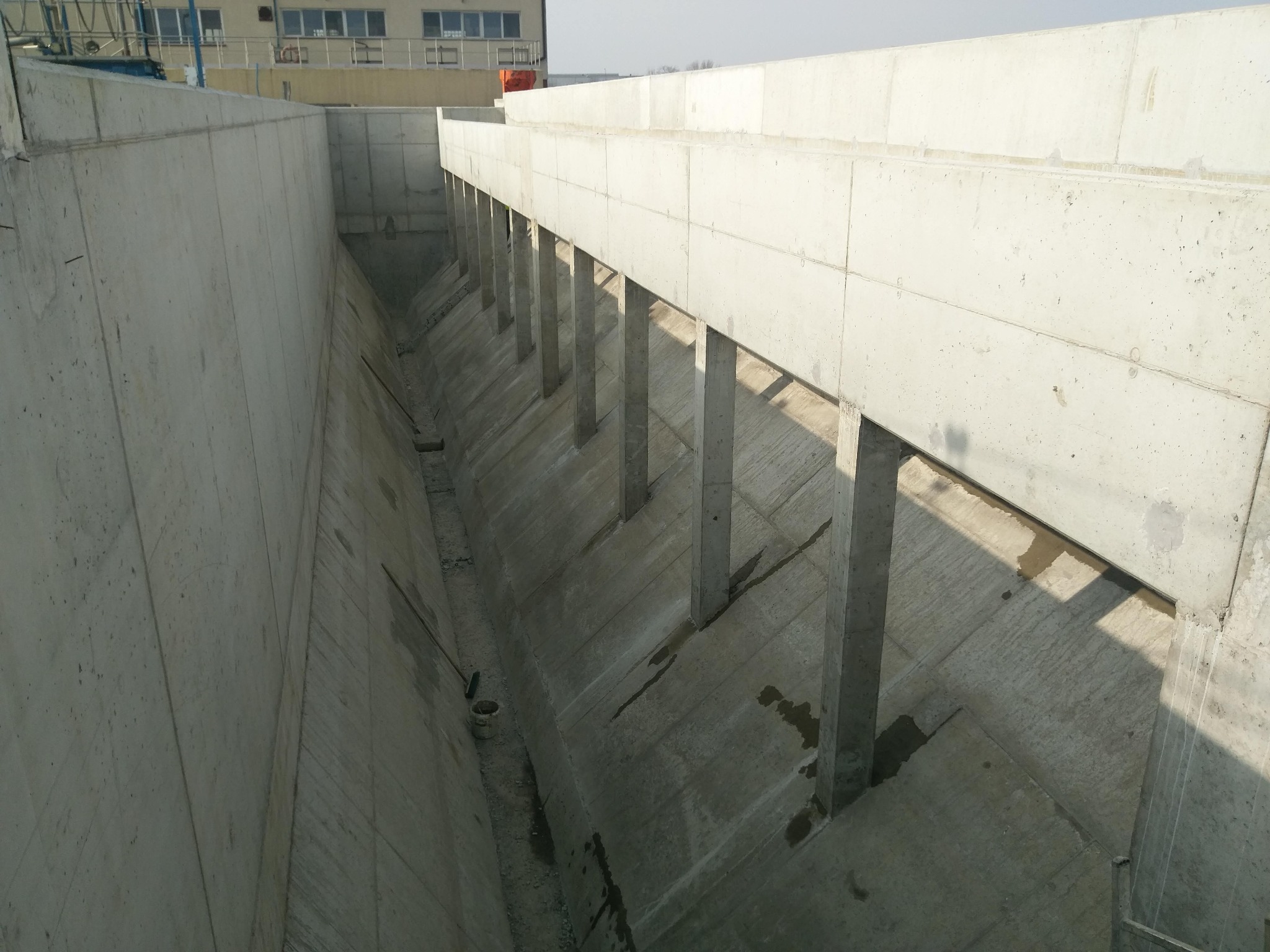 Betonowy kanał z podporami, widoczne ślady wilgoci i narzędzia budowlane, budynek w tle.