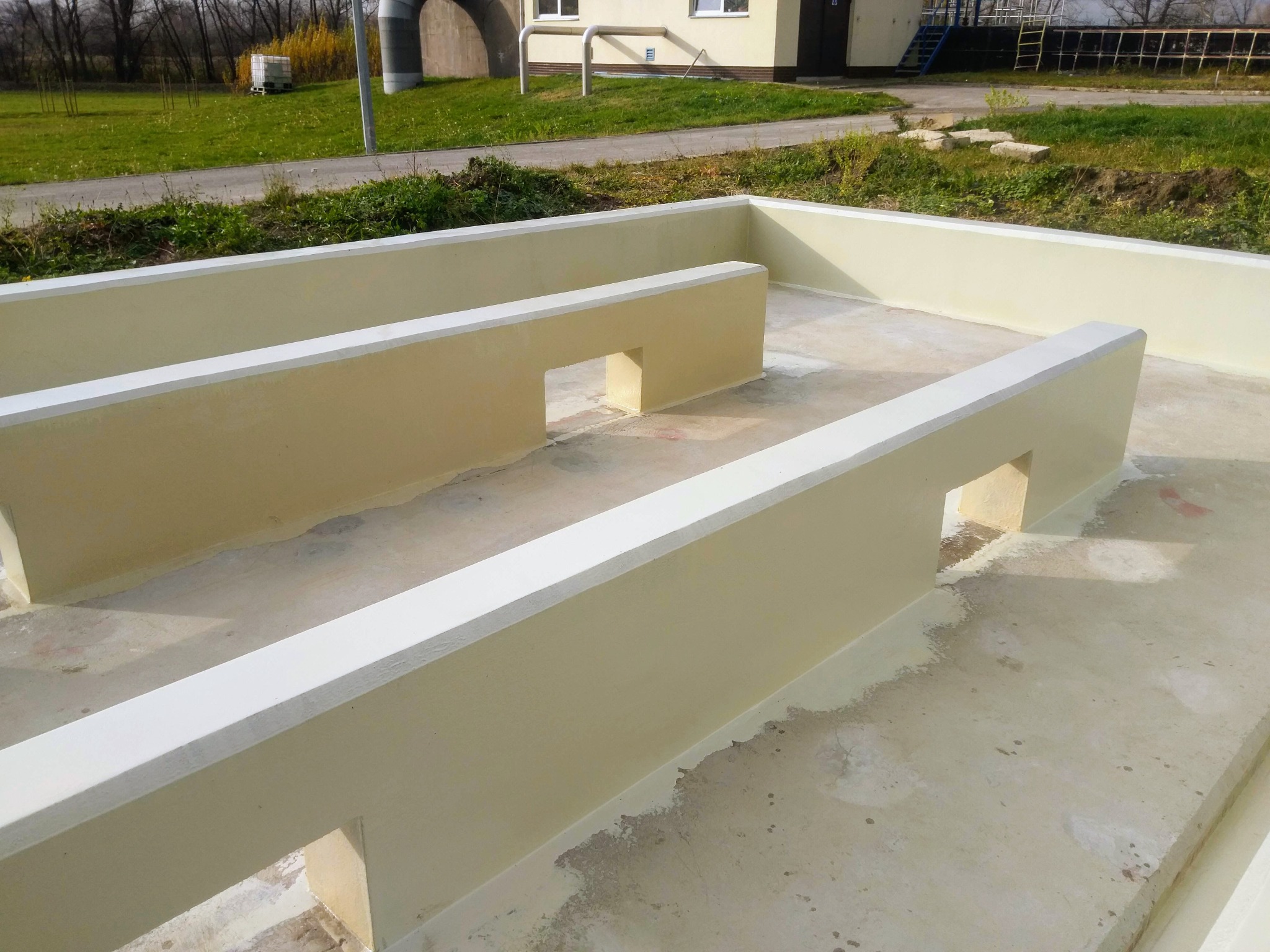 Betonowa konstrukcja z niskimi ściankami działowymi, świeżo pomalowana na beżowo z białymi krawędziami, widoczna na tle budynku i zieleni.