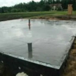 Wylana, gładka płyta fundamentowa z wystającymi rurami kanalizacyjnymi, otoczona czarną izolacją termiczną, na tle zieleni i drzew.