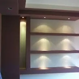 Ściana z półkami z płyty gipsowo-kartonowej, oświetlona punktowymi lampkami, z dekoracyjnym elementem na suficie w kolorze bordowym, widoczne niedoskonałości w jakości zdjęcia.