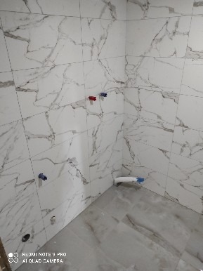 Wnętrze łazienki w trakcie remontu, z marmurowymi płytkami na ścianach i podłodze, widoczne przyłącza hydrauliczne z niebieskimi i czerwonymi zaślepkami, oraz fragment rury kanalizacyjnej w rogu.