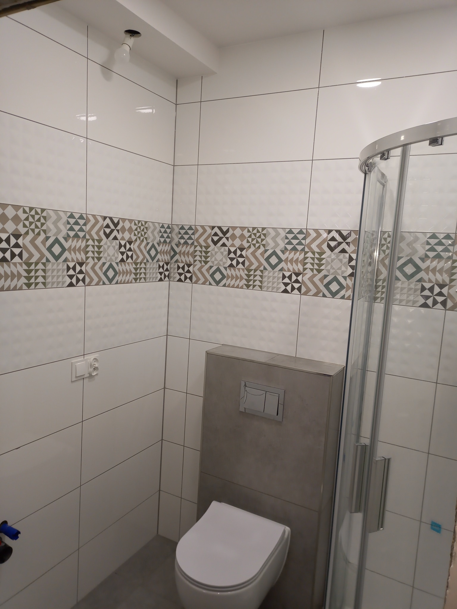 Nowoczesna łazienka z białymi płytkami, dekoracyjnym pasem z geometrycznym wzorem, podwieszaną toaletą i kabiną prysznicową z przezroczystego szkła.