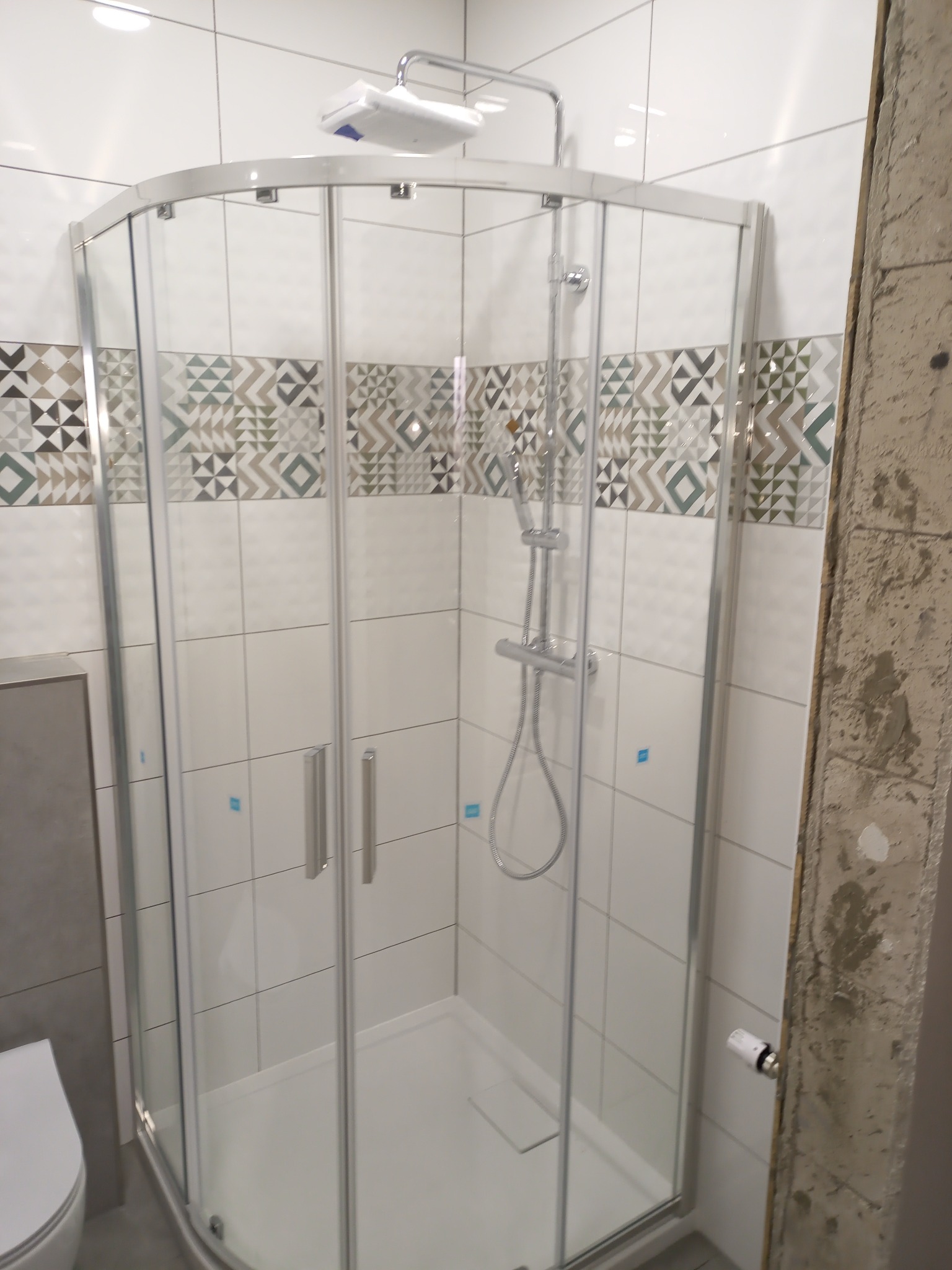 Kabina prysznicowa narożna z dwuskrzydłowymi drzwiami przesuwnymi, zamontowana na białym brodziku, z widoczną baterią prysznicową i deszczownicą, na ścianach białe płytki z dekoracyjnym pasem...