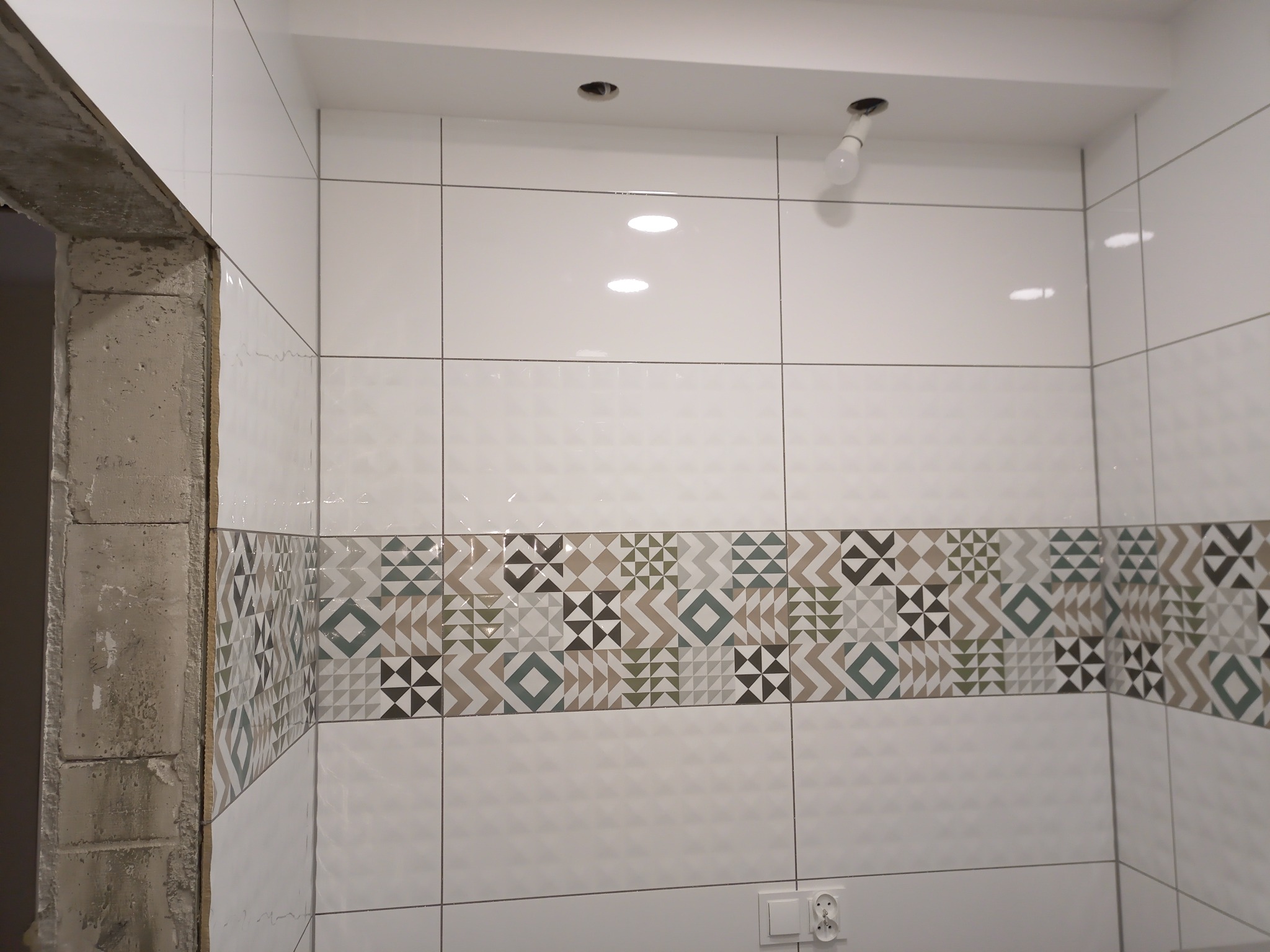 Ściana łazienki wyłożona białymi płytkami z dekoracyjnym pasem mozaiki geometrycznej, widoczne gniazdko elektryczne i surowy fragment ściany przy wejściu.