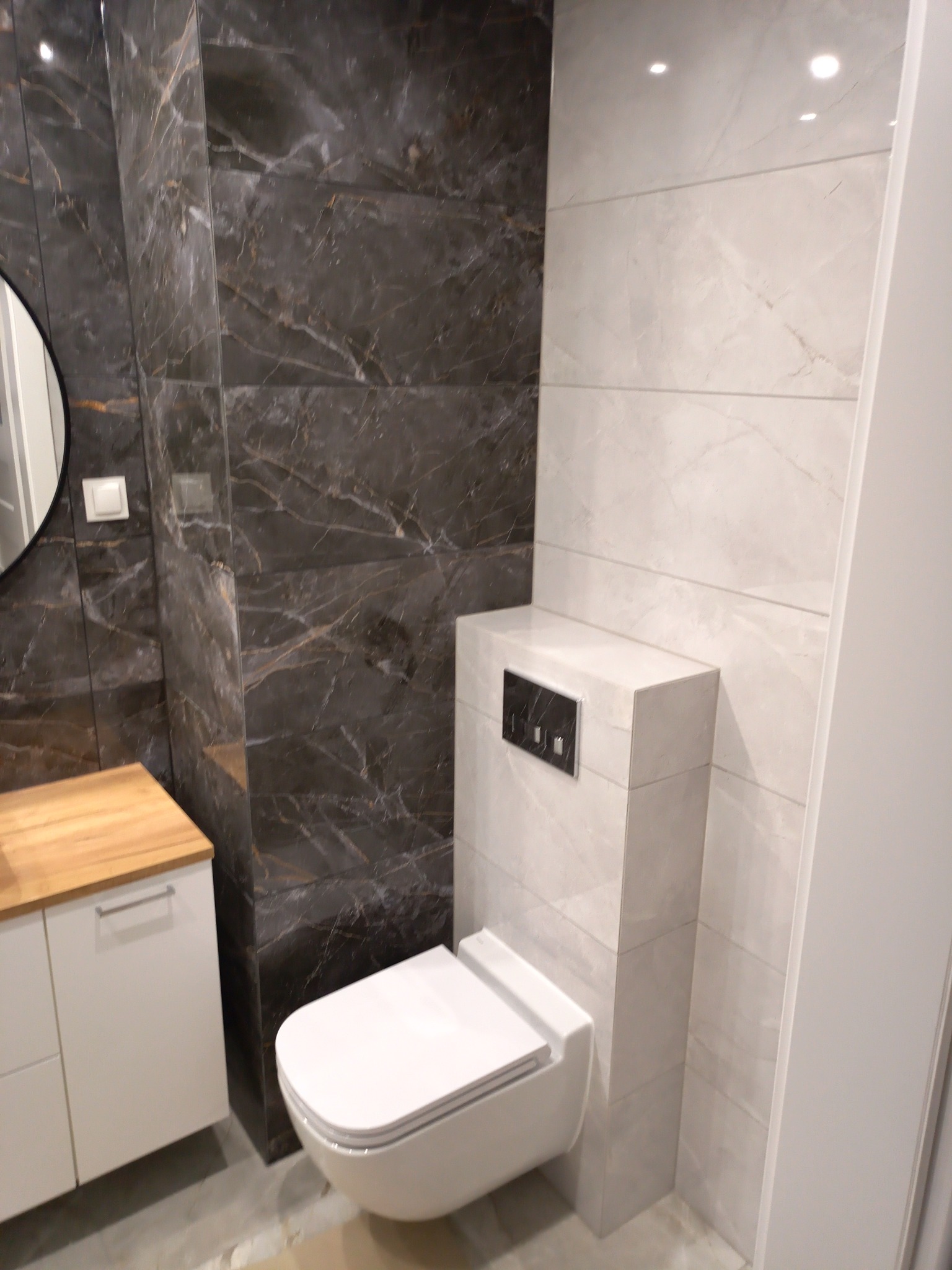 Nowoczesna łazienka z podwieszaną toaletą, zabudową geberit w płytkach imitujących marmur oraz szafką z drewnianym blatem, ściany wyłożone jasnymi i ciemnymi płytkami.