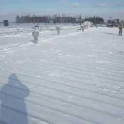 Pracownik w odblaskowej kamizelce usuwa śnieg z płaskiego dachu budynku w słoneczny dzień, widoczny cień osoby robiącej zdjęcie.