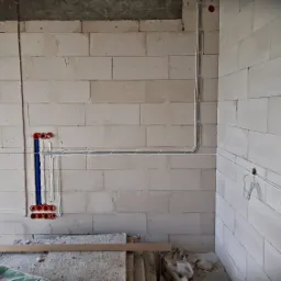Ściana z betonu komórkowego w trakcie prac instalacyjnych, widoczne rury miedziane i peszle z przewodami w kolorze niebieskim oraz pomarańczowe puszki instalacyjne pod gniazdka i włączniki.