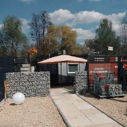 Ekspozycja systemów ogrodzeniowych: gabiony z kamieniami, panele i siatki, z siedzibą firmy w tle i ścieżką z jasnych płyt.