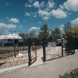 Otwarta dwuskrzydłowa brama w ciemnej ramie, część ekspozycji z różnymi typami ogrodzeń panelowych i siatkowych na żwirowym podłożu, w tle biały namiot ekspozycyjny i błękitne niebo z chmurami.