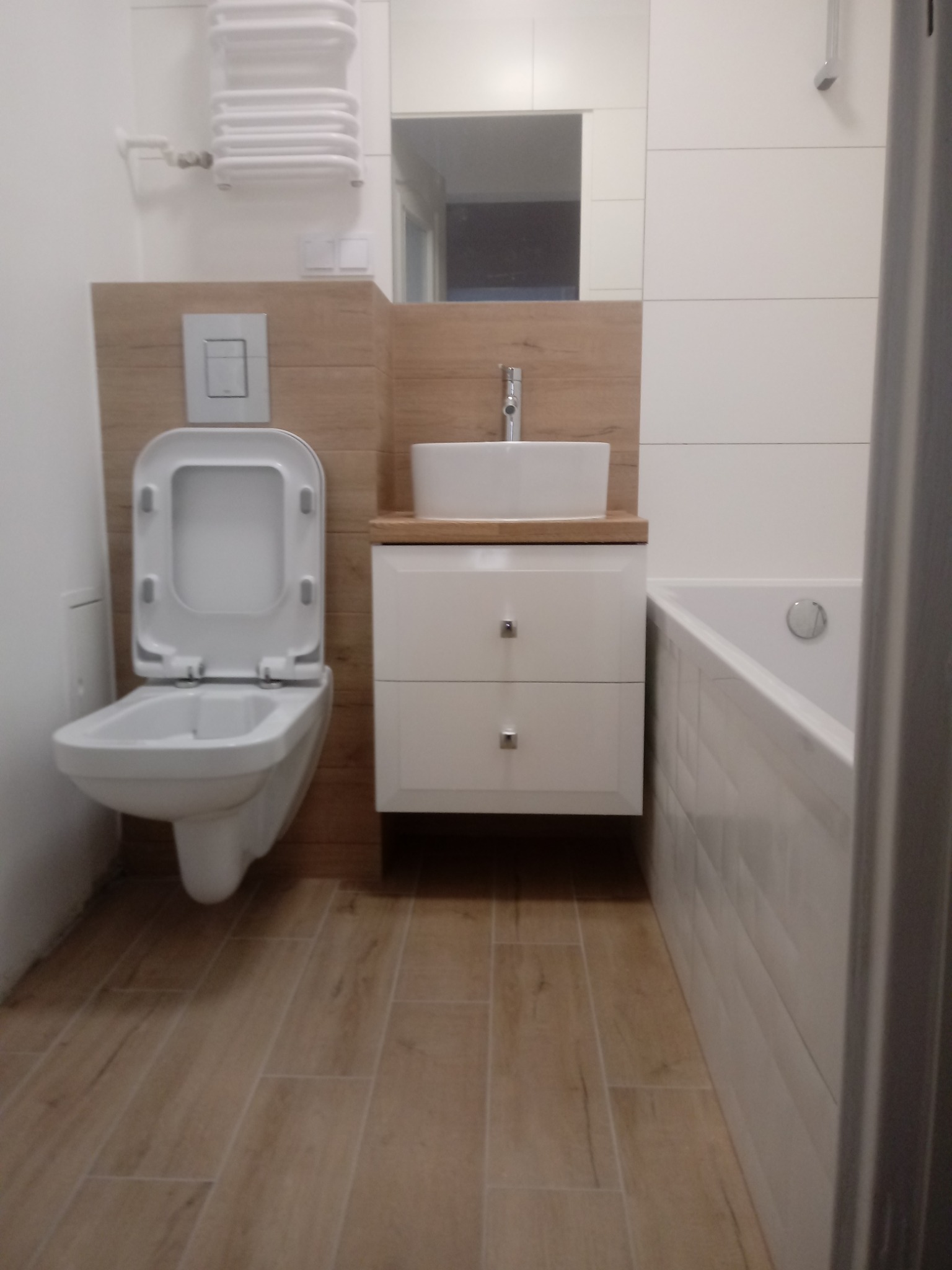 Nowoczesna łazienka z toaletą z otwartą deską, białą umywalką na szafce z drewnianym blatem, grzejnikiem drabinkowym i płytkami imitującymi drewno na podłodze.