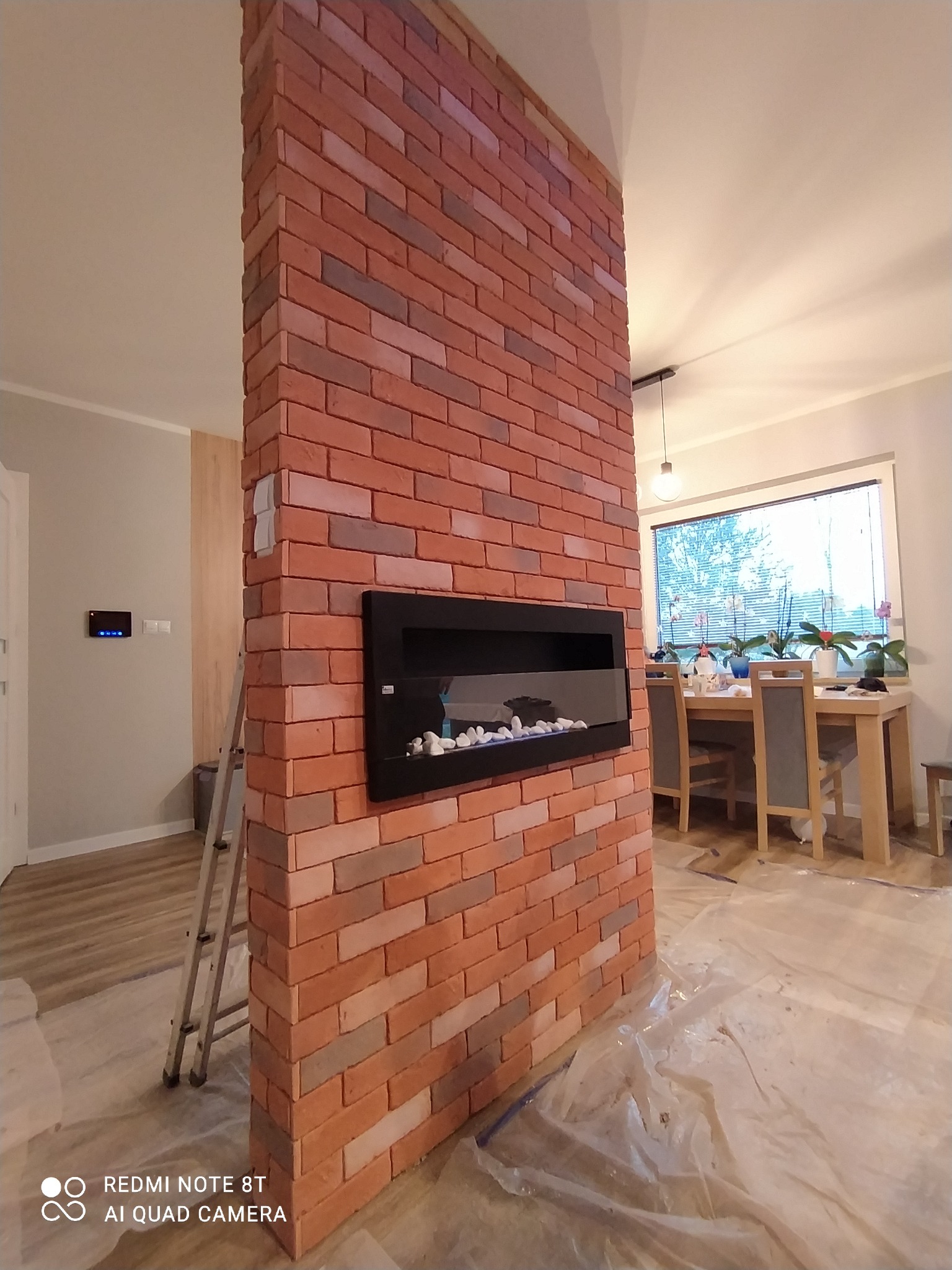 Ścianka działowa z cegły klinkierowej z wbudowanym biokominkiem elektrycznym, widoczna drabina malarska, zabezpieczona folią podłoga w trakcie prac wykończeniowych.