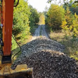 Pomarańczowa koparka Doosan częściowo widoczna z lewej strony, na tle usypanej drogi z kamieni w lesie, widoczne ślady opon.