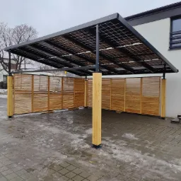 Zadaszenie w nowoczesnym stylu z panelami słonecznymi i drewnianą osłoną, wsparte na drewnianych słupach, na tle budynku mieszkalnego.