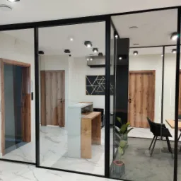 Przeszklone ściany działowe z czarnymi profilami w nowoczesnym biurze, widoczne drzwi z drewnianą okleiną, marmurowa podłoga i minimalistyczne meble.