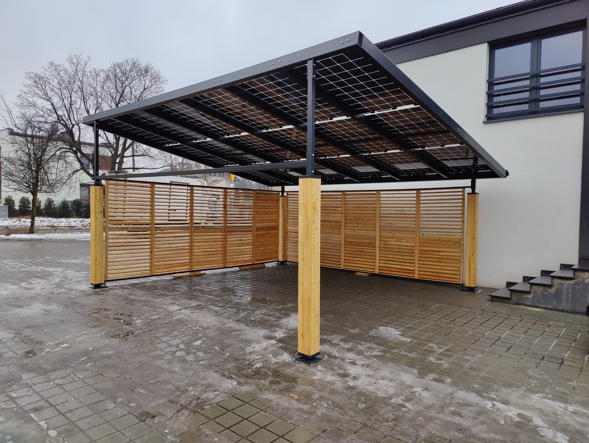 Zadaszenie w nowoczesnym stylu z panelami słonecznymi i drewnianą osłoną, wsparte na drewnianych słupach, na tle budynku mieszkalnego.