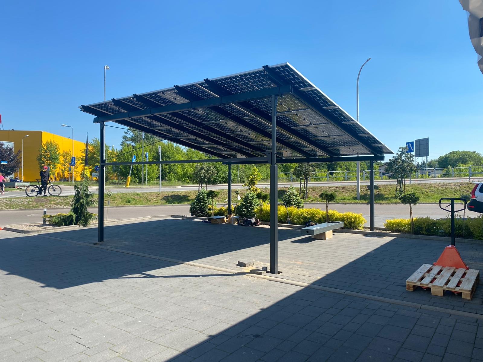 Zadaszenie parkingu z panelami słonecznymi, widoczny wózek paletowy i żółty budynek w tle.