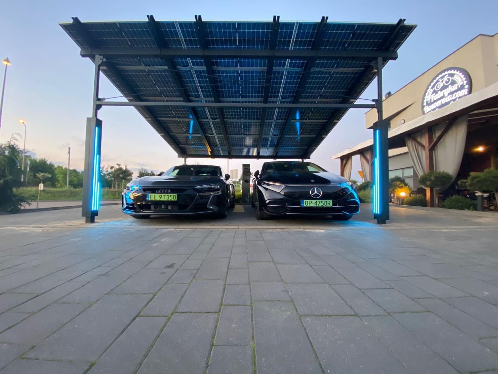 Dwa czarne samochody elektryczne Audi i Mercedes zaparkowane pod zadaszeniem z paneli słonecznych z niebieskim oświetleniem LED.