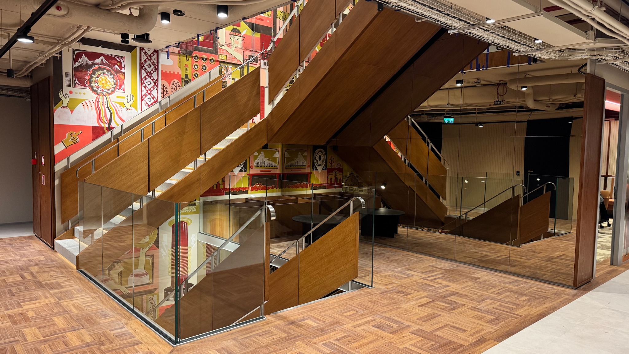 Nowoczesne schody z drewnianymi stopniami i szklanymi balustradami w przestrzeni komercyjnej z artystycznymi malowidłami na ścianach i parkietem w jodełkę.