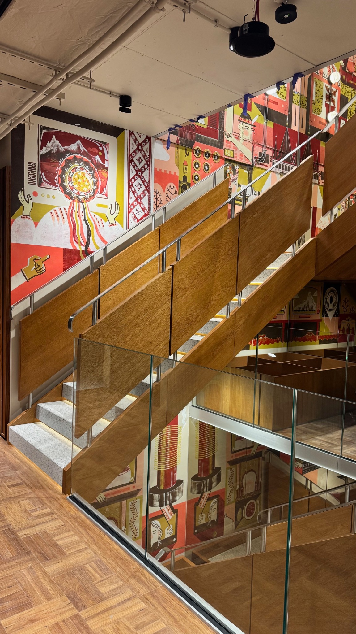 Nowoczesne schody z drewnianymi panelami i szklanymi balustradami, ozdobione kolorowymi muralami na ścianach. Widok z dołu, podkreślający geometrię i artystyczny charakter.