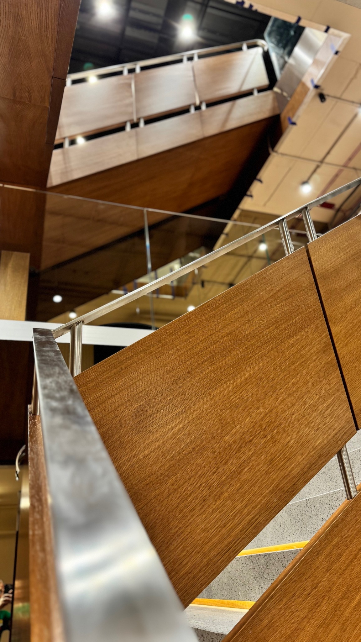 Wielopoziomowe schody z drewnianymi panelami i metalową balustradą, widziane z dołu pod nietypowym kątem, z widocznymi elementami oświetlenia sufitowego.