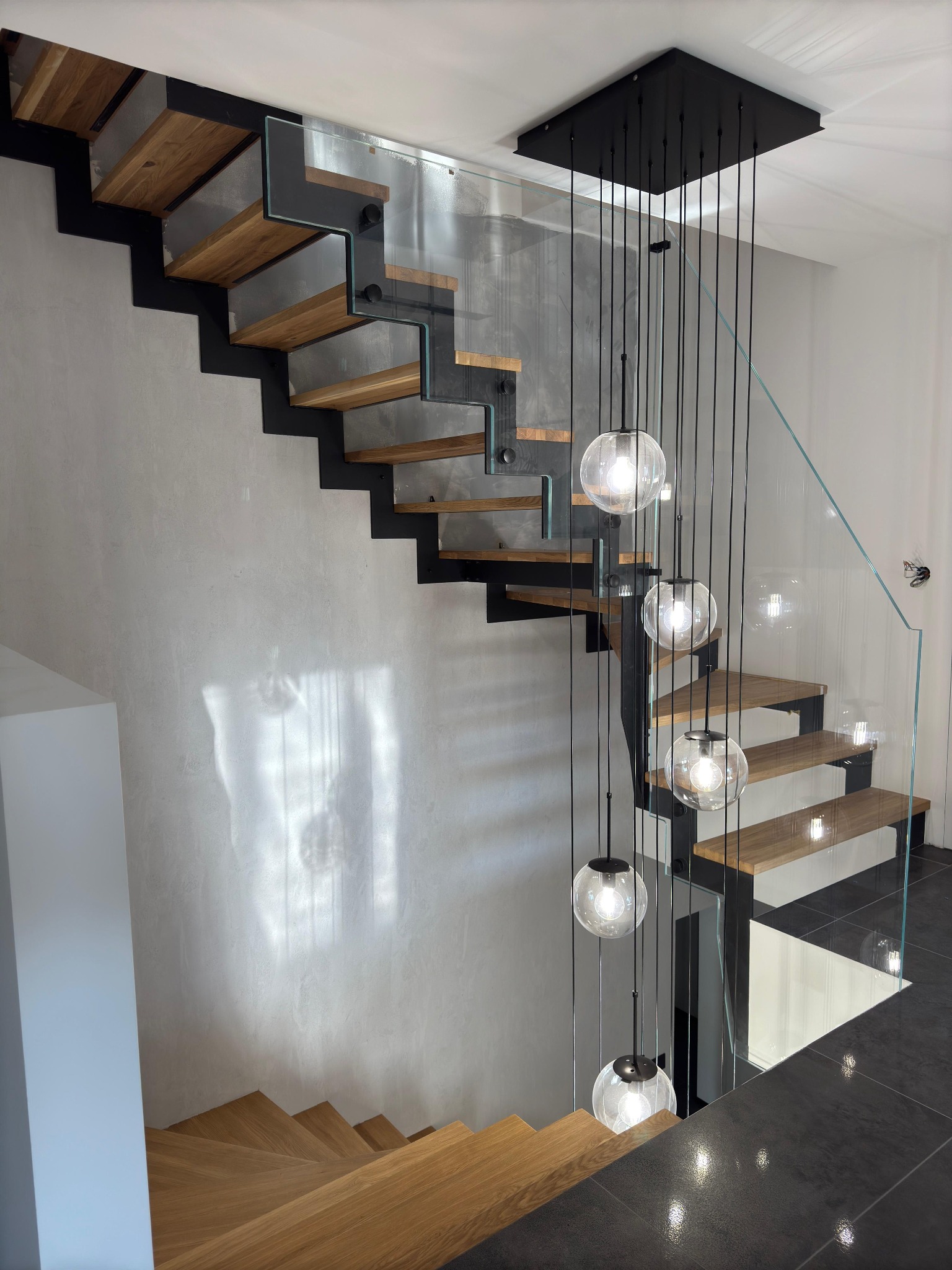 Nowoczesne schody z drewnianymi stopniami, czarną metalową konstrukcją i szklaną balustradą. Nad schodami zwisają szklane kule oświetleniowe na czarnych kablach.