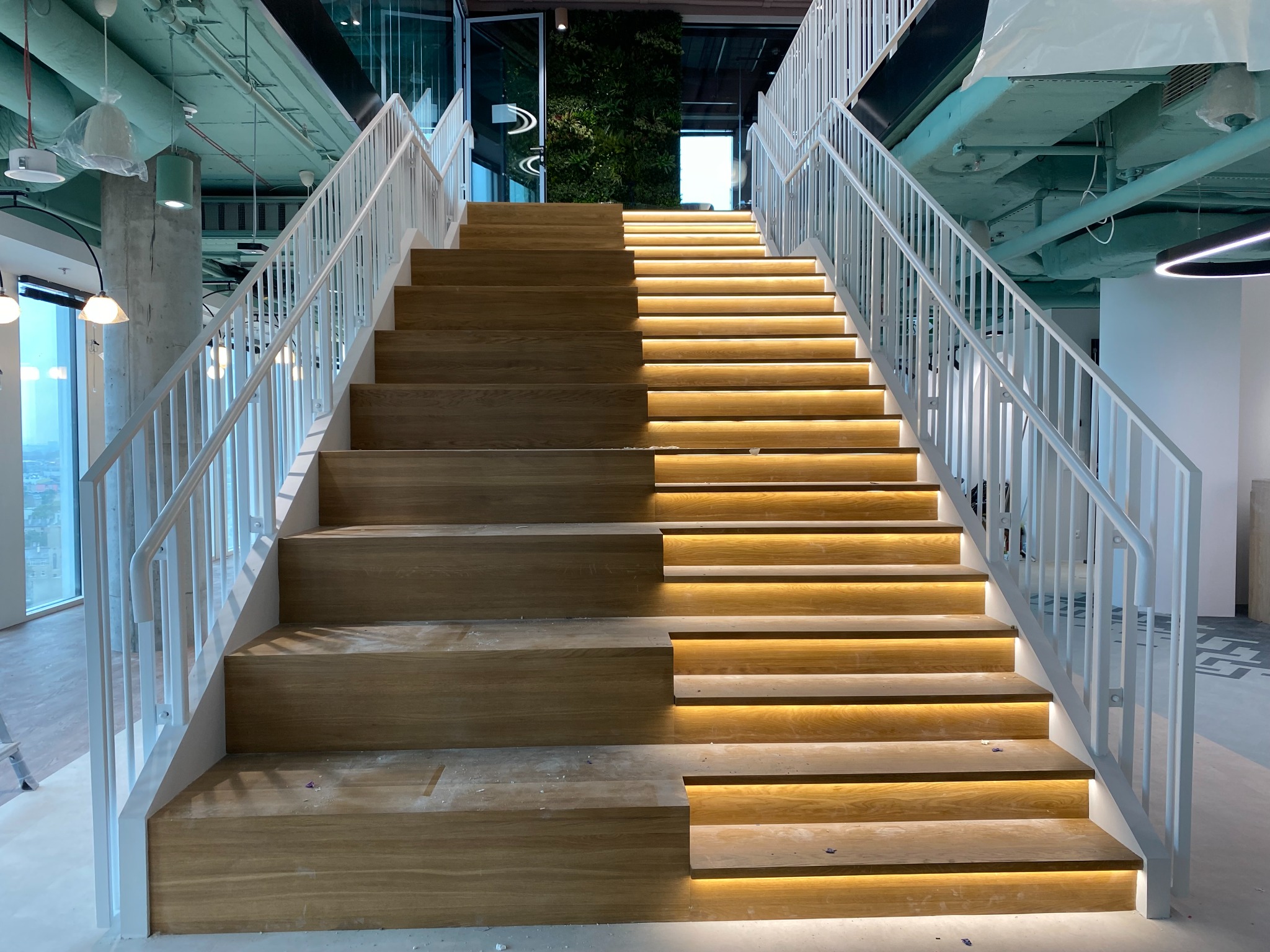 Nowoczesne schody z drewnianymi stopniami i białymi balustradami, podświetlone od spodu, prowadzące do zielonej ściany w tle. Widok z dołu, jasne oświetlenie.