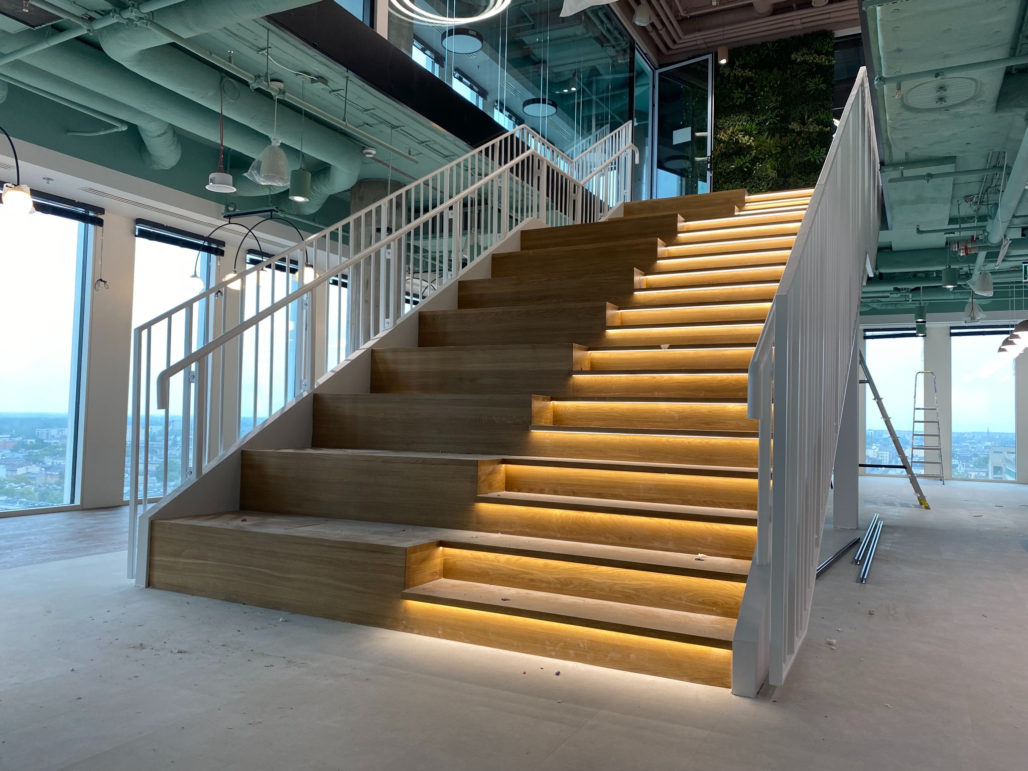 Nowoczesne schody z drewnianymi stopniami podświetlonymi LED, białe balustrady, widok na miasto z okien. Elementy wykończeniowe na podłodze sugerują trwające prace.