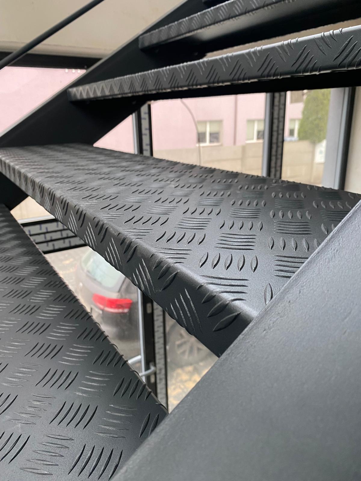 Czarne schody metalowe z blachy ryflowanej, widok z dołu, z oknem w tle. Detal stopni z antypoślizgową fakturą.