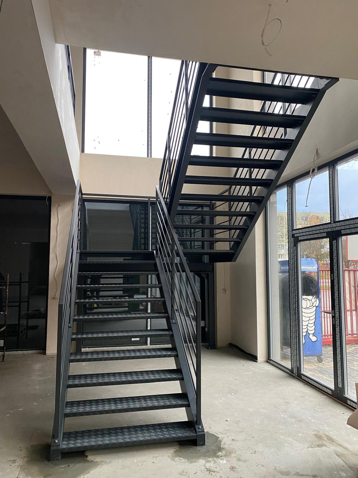 Nowoczesne, dwubiegowe schody metalowe z balustradą, z perforowanymi stopniami, w jasnym wnętrzu z dużymi oknami. Widok z dołu, podkreślający konstrukcję.