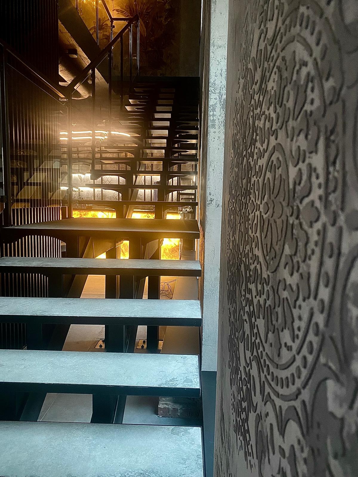 Metalowe schody z podświetleniem, widok z dołu, z dekoracyjną ścianą po prawej. Elementy konstrukcyjne schodów są czarne, stopnie szare. Nietypowe oświetlenie.