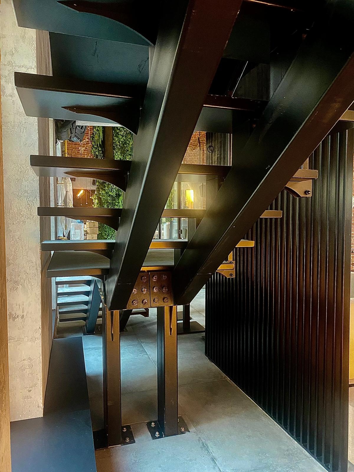 Nowoczesne schody metalowe z czarnymi stopniami i konstrukcją, widoczne detale mocowań śrubowych, w tle elementy dekoracyjne wnętrza.