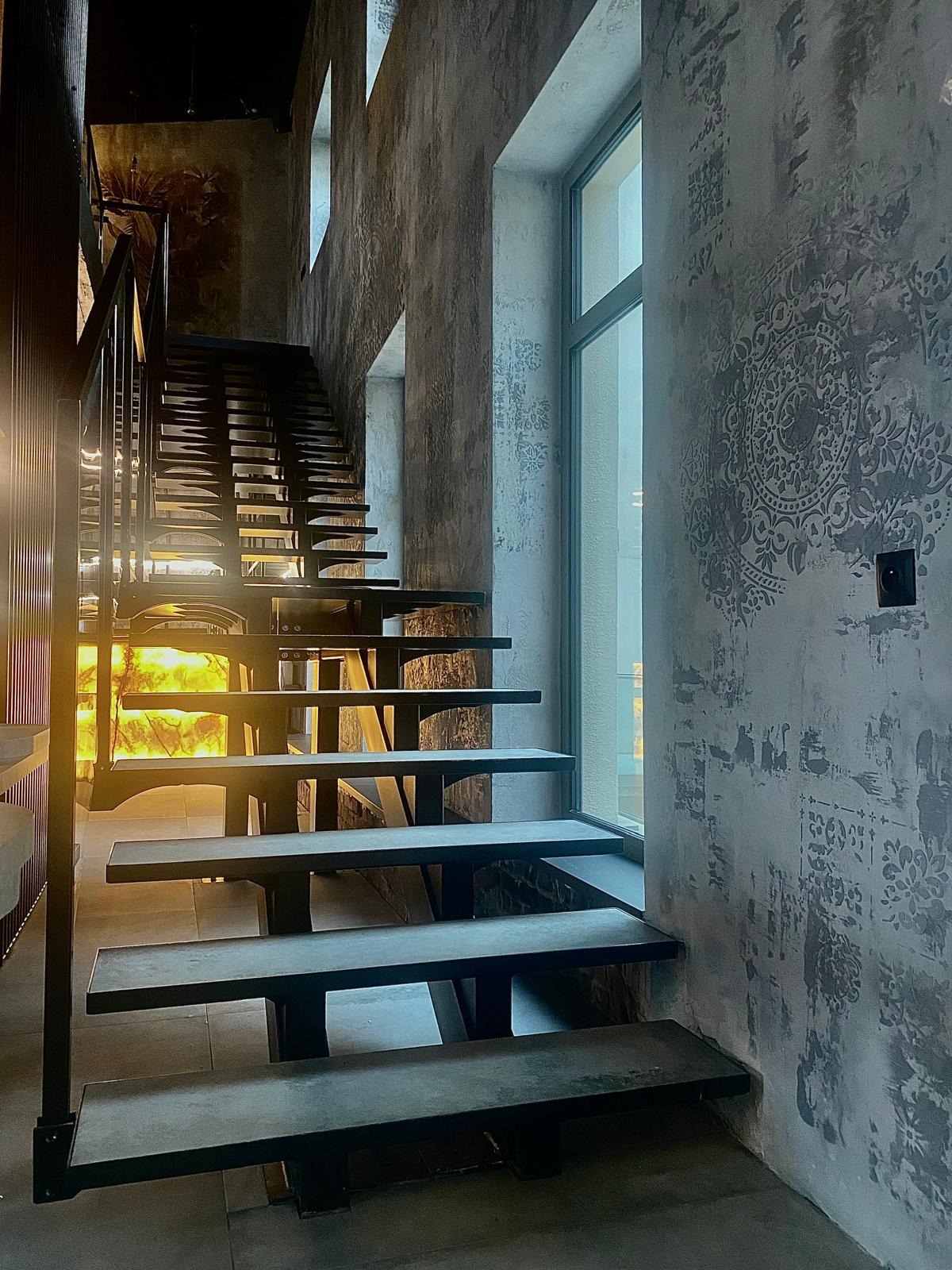 Nowoczesne schody metalowe z czarną balustradą, oświetlone od dołu, na tle ściany z dekoracyjnym wzorem i oknem. Minimalistyczny design, surowy styl.