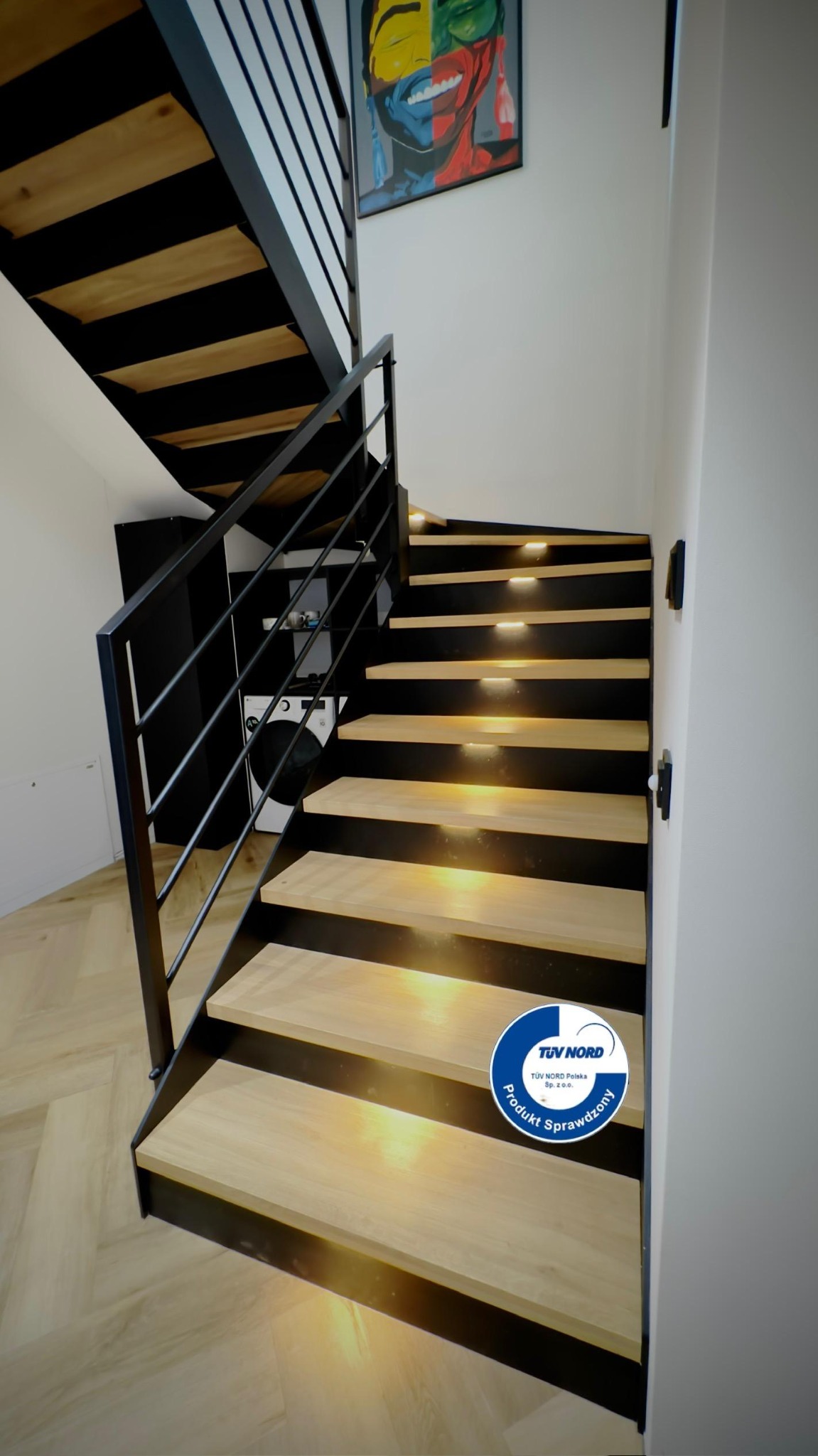 Nowoczesne schody z drewnianymi stopniami i czarną metalową balustradą, oświetlone od spodu, z widocznym certyfikatem jakości TÜV NORD na jednym ze stopni.