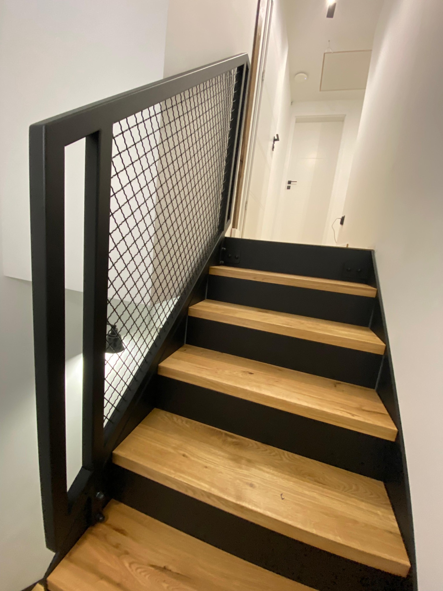Nowoczesne schody z drewnianymi stopniami i czarną, metalową balustradą z siatką. Widok z dołu, oświetlenie górne, minimalistyczny design.
