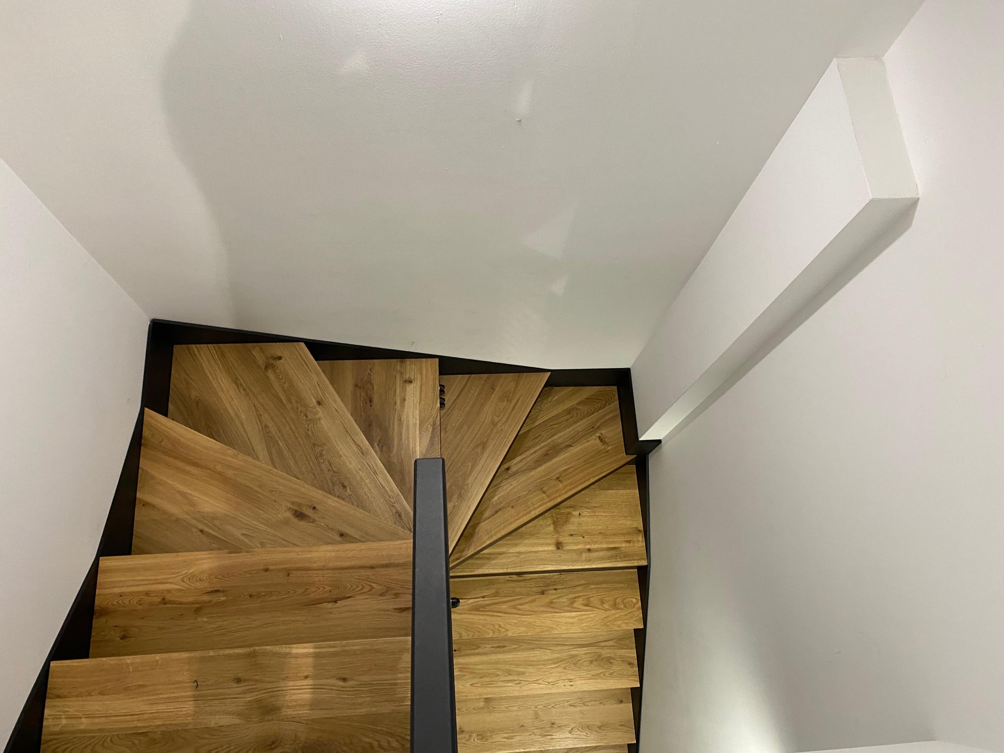 Spiralne schody z drewnianymi stopniami i czarną metalową balustradą, widziane z góry, w jasnym wnętrzu. Minimalistyczny design.