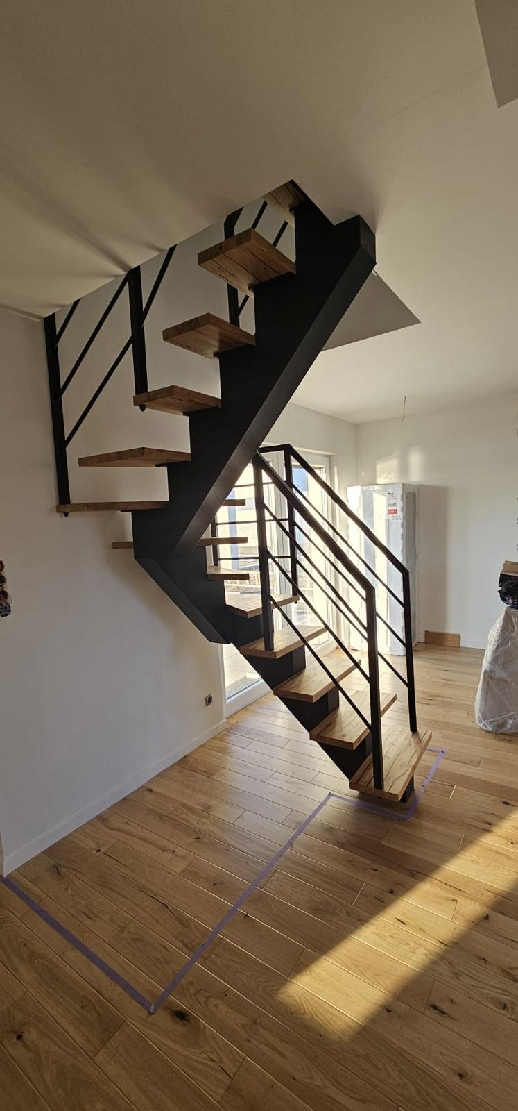 Nowoczesne schody metalowe z drewnianymi stopniami, minimalistyczny design, jasne wnętrze z parkietem. Widoczne metalowe barierki i konstrukcja schodów.
