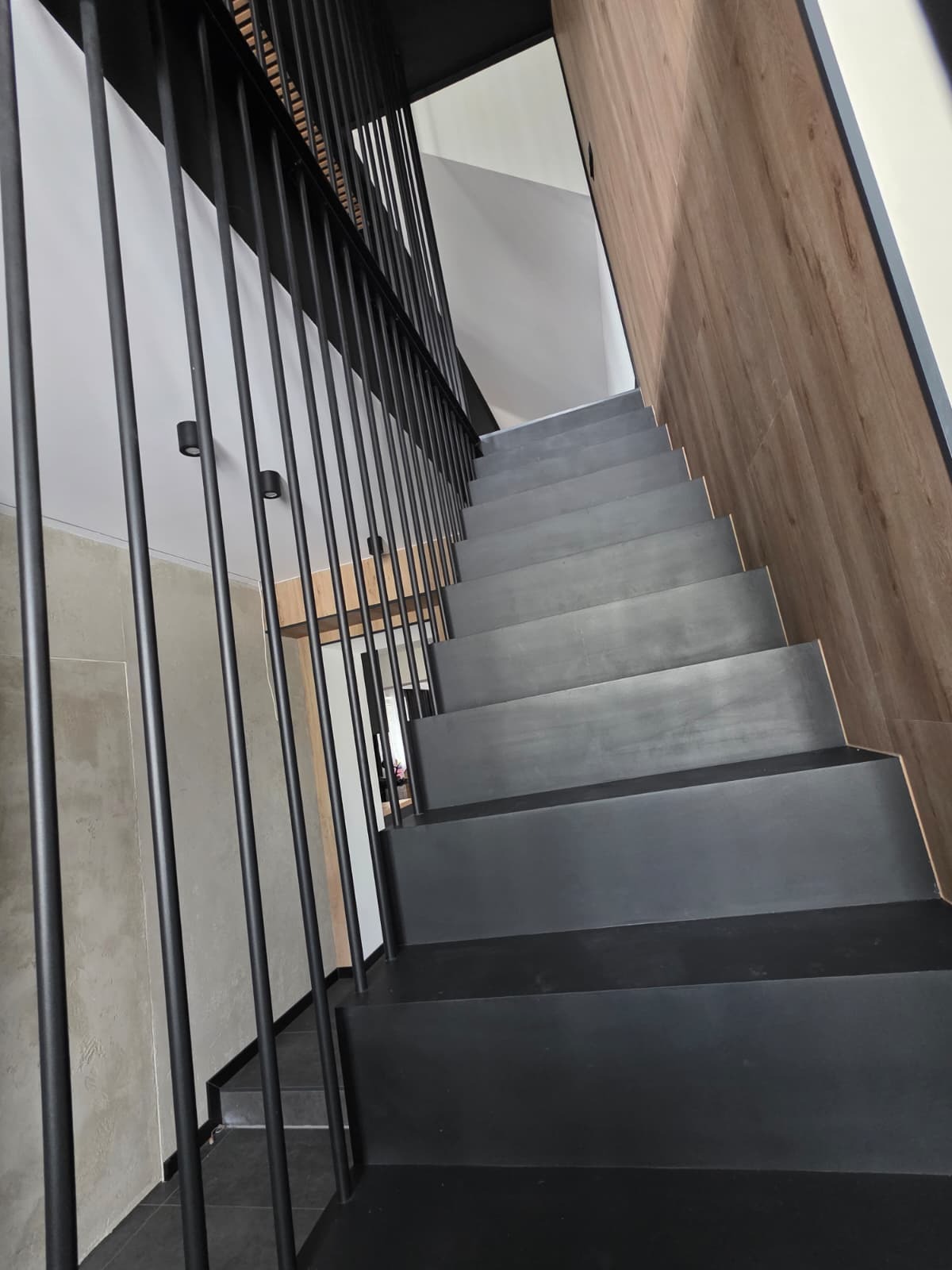 Nowoczesne, czarne schody metalowe z minimalistyczną balustradą z pionowych prętów, widok z dołu, z drewnianą okładziną ściany po prawej stronie.