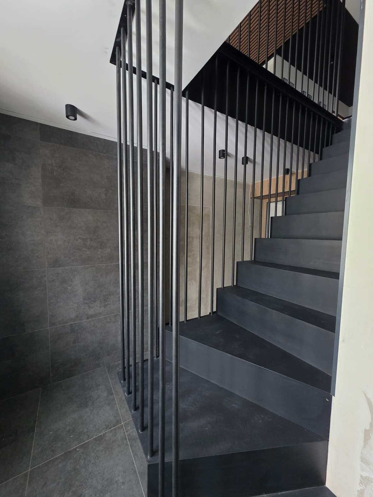 Nowoczesne, czarne schody metalowe z minimalistyczną balustradą z pionowych prętów. Widok z dołu, podkreślający geometrię i surowy charakter wnętrza.