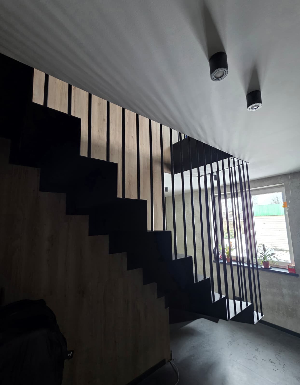 Nowoczesne schody z czarnymi, pionowymi balustradami, drewnopodobne wykończenie stopni, oświetlenie sufitowe punktowe, widok z dołu, minimalistyczny design.
