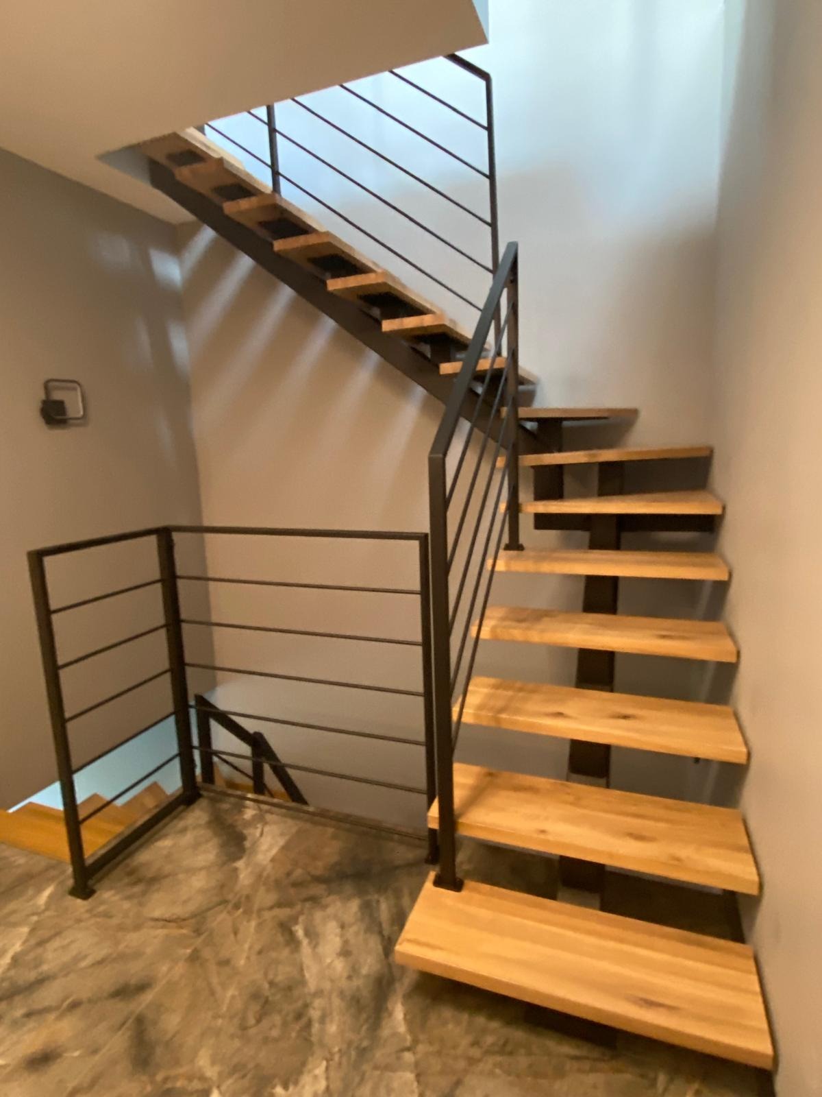 Nowoczesne schody z drewnianymi stopniami i czarną, minimalistyczną balustradą. Widok z dołu, ukazujący konstrukcję i detale wykończenia.