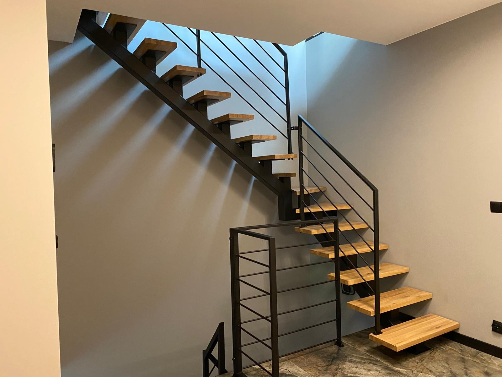 Nowoczesne schody z drewnianymi stopniami i czarną metalową konstrukcją, minimalistyczny design, szare ściany, widok z dołu, oświetlenie górne.