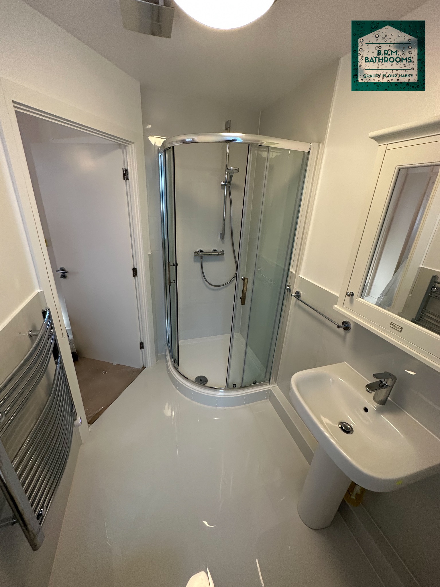 Jasna łazienka po remoncie z kabiną prysznicową z zaokrąglonymi drzwiami, umywalką na postumencie, grzejnikiem drabinkowym i otwartymi drzwiami do pomieszczenia z surową podłogą.