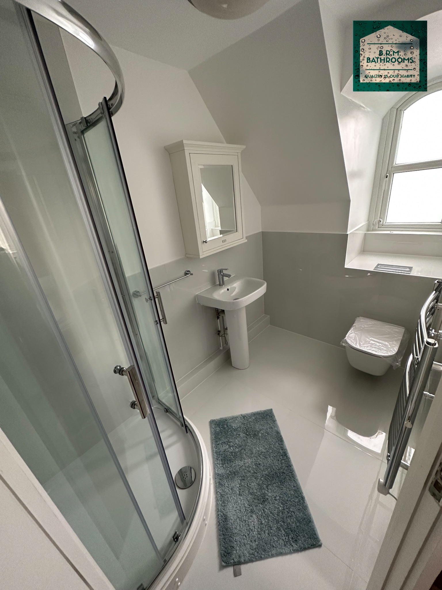 Jasna łazienka na poddaszu z kabiną prysznicową, umywalką na postumencie, toaletą w folii ochronnej i szarą matą podłogową, oświetlona naturalnym światłem z okna dachowego.