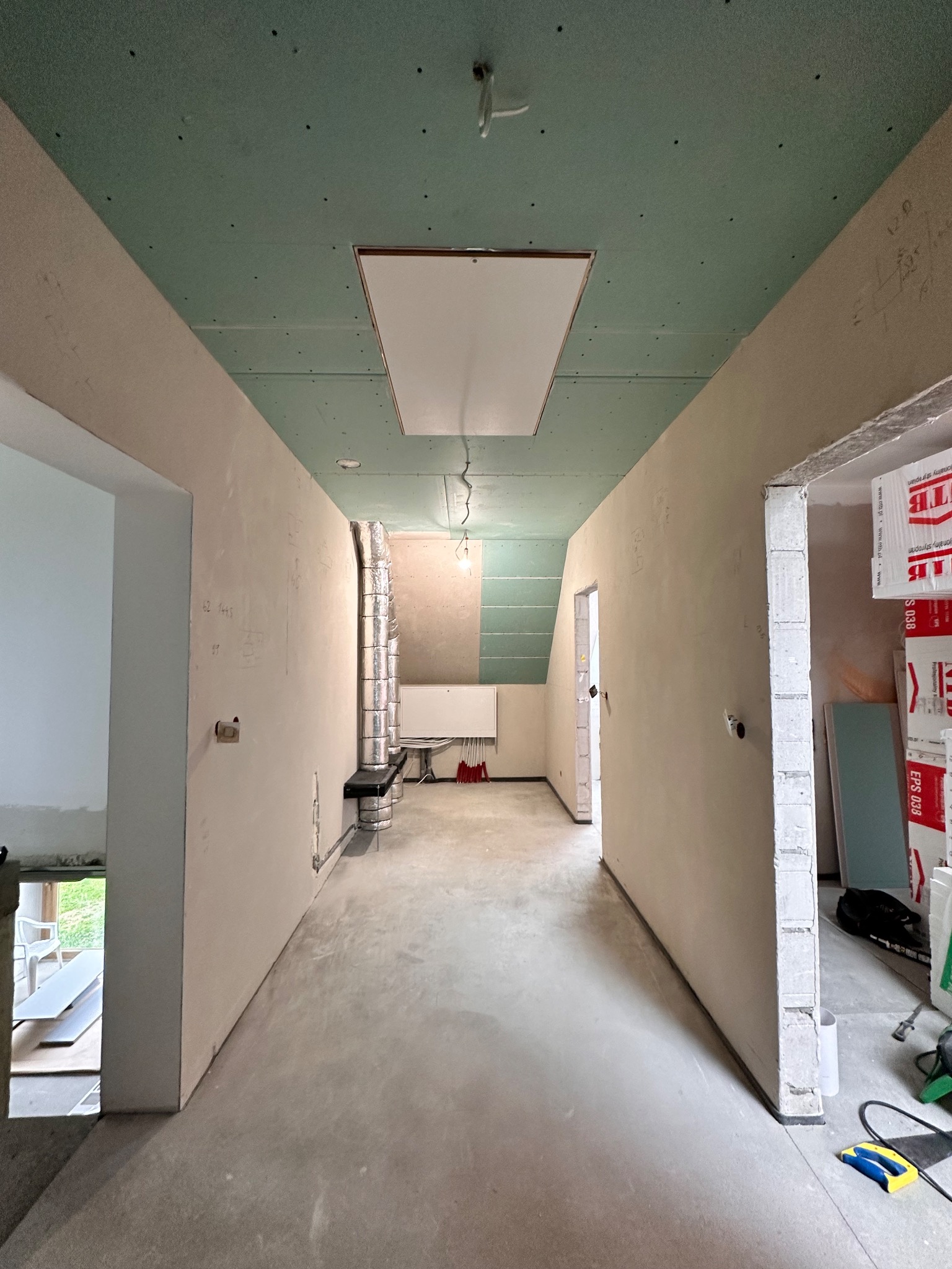 Wnętrze w trakcie prac wykończeniowych: surowy beton na podłodze, ściany częściowo pokryte gładzią z widocznymi notatkami, sufit z płyt gipsowo-kartonowych w kolorze zielonym, widoczne instalacje...