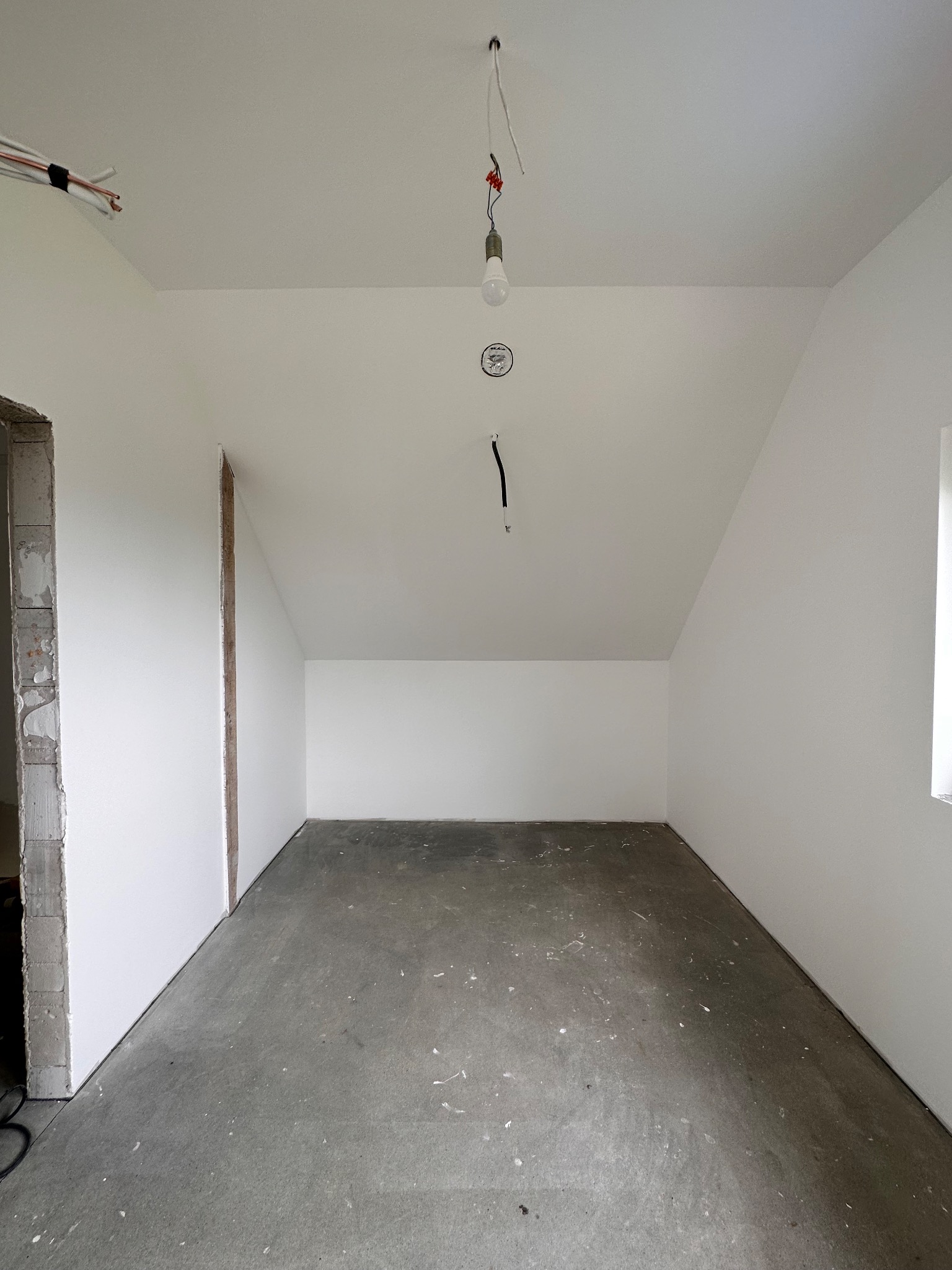Surowe wnętrze poddasza ze świeżo pomalowanymi białymi ścianami, betonową podłogą z resztkami farby, widocznymi przewodami elektrycznymi i surowym otworem drzwiowym.